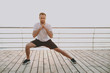 Full length portrait of handsome young bearded athletic man guy 20s in white t-shirt posing training doing stretching lunge exercising for legs looking camera at sunrise over the sea outdoors.