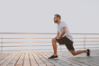 Side view full length portrait attractive young bearded athletic man guy 20s in white t-shirt posing training doing stretching lunge exercising for legs looking aside at sunrise over the sea outdoors.