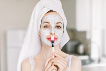 Close up of beautiful young woman with bath towel on her head, applying lifting hydrating moisturizing facial mask to her face at modern kitchen, holding cosmetic brush. Skin care treatment at home