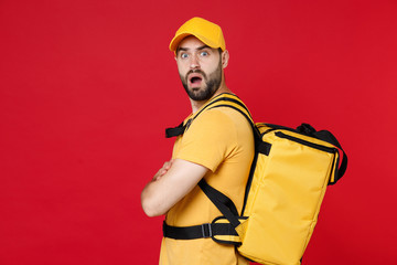 Wall Mural - Side view delivery man 20s in yellow cap t-shirt thermal bag backpack with food isolated on red background studio Guy male employee work courier Service quarantine coronavirus covid-19 virus concept