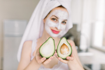 Young smiling attractive woman holding two halves of avocado, with anti aging facial cream mask, posing over blurred home kitchen background, showing avocado to camera. Focus on avocado