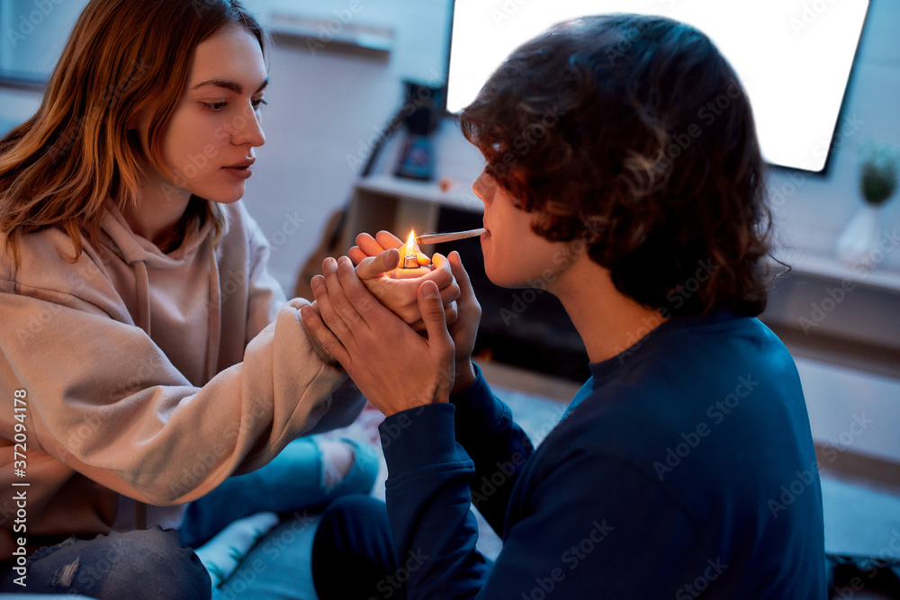 Young girl lighting cannabis cigarette for her boyfriend while sitting on  the couch at home. Young couple smoking marijuana joint and relaxing Stock  Photo