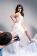 © danmorgan12 - Portrait of Man Taking Pictures of Brunette Caucasian Female In White Wedding Dress. Posing With Leg On High Prop Against Gray