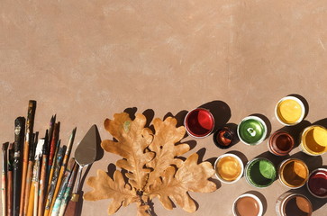 Drawing tools on artist table and fall autumn leaf top view on beige paint textured background. Copy space