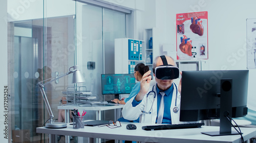 Senior physician using VR glasses in modern private clinic to study diseases in virtual space and modern technology. In the background modern clinic with glass walls and patients with doctors in