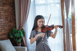 © Odua Images - asian girl in the dark gray dress plays the violin smiling while looking at the camera in the room against the window curtain