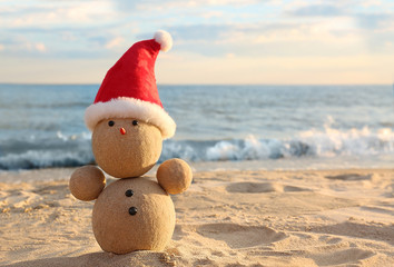 Snowman made of sand with Santa hat on beach near sea, space for text. Christmas vacation