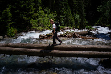 Hiker man hiking crossing river walking in balance on fallen tree trunk