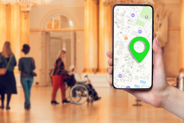 A females hand holds a smartphone with an online map on the screen. In the background, a group of people and a man in a wheelchair in a Museum, in a blur. Concept of online applications and navigation