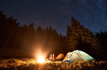 Wall Mural - young couple hikers sitting by bright burning bonfire near two tents, enjoying beautiful camping nig