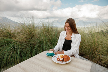 Canvas Print - Young beautiful woman smiles enjoys hot cpoffee  on the terrace of a cafe in the mountain