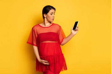 Wall Mural - upset pregnant womani n red tunic holding smartphone with blank screen while touching belly on yellow