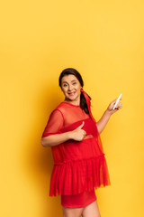 Wall Mural - young pregnant woman in red tunic showing thumb up while holding pregnancy test on yellow