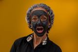 Fototapeta  - Brazilian woman with surprised expression and gray clay mask. Concept: beauty, face care, facelift, wellness, skin care. Shot with orange background