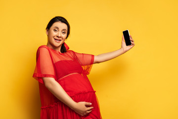 Wall Mural - brunette pregnant woman in red tunic taking selfie on smartphone while looking at camera on yellow