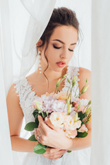 Wall Mural - Selective focus of bride in wedding dress holding bouquet near white curtains