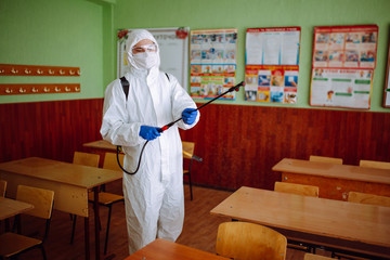 Wall Mural - A man in protective antibacterial suit cleaning up the classroom with a spray with sanitizing liquid. Professional sanitary worker disinfects the auditorium with special equipment. Healthcare concept.