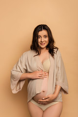 Wall Mural - young pregnant woman in shirt and lingerie touching belly and looking at camera on beige