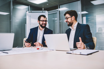 Two office workers in business meeting sitting at desk using computer for business training, professionals smiling and using modern electronics devices during conversation about successful project