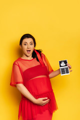 Wall Mural - pregnant woman in red tunic touching belly while showing ultrasound scan on yellow