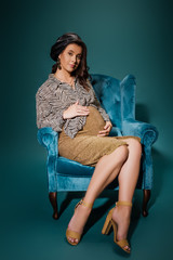 Wall Mural - pregnant woman in stylish clothes touching tummy while sitting in velour armchair on dark blue