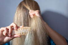 Blonde Combs Dry Hair With A Wooden Comb. On A Gray Background. Concept Of Dry Female Blonde Hair. Damaged Hair. Breaking Damaging Hair
