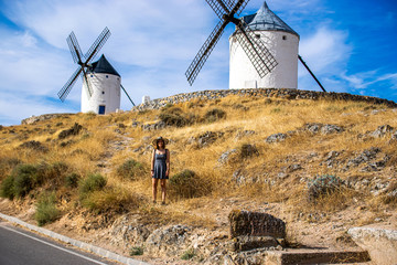 Photo of an attractive woman standing next to some beautiful and historic windmills located in Consuegra, Toledo, Spain during a sunny day of summer in a natural place. 