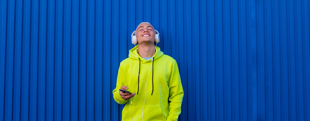 close up and portrait of teenager young man listening music and having fun with his phone and wearing headphones - blue background wall - cheerful and happy millennial hearing music ad vibing