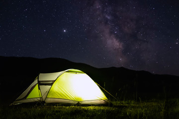 Illuminated green tent with mountains silhouette and many stars background. Hiking, tourism concept. Adventure travel. Outdoor landscape. Night sky, stars background.