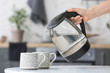 Woman pouring hot water from electric kettle into cup on kitchen table