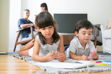 Focused cute little brother and sister lying on floor and drawing in living room while parents sitting together in background. Childhood or kids creative hobby concept