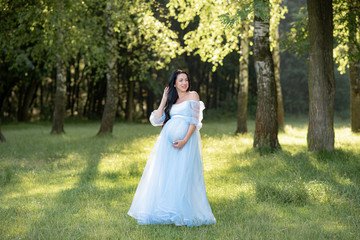  Pregnant woman posing in a blue dress on a background of green trees.