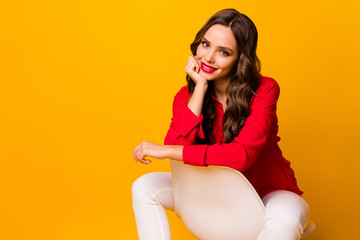 Poster - Photo of attractive business lady toothy smiling sit cosy chair between legs lean chin arm cheerful wear red shirt white trousers isolated yellow vivid color background