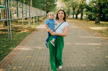 Beautiful caucasian woman with dark hair in white t-shirt and long green skirt hugs and rejoices with her lovely son with short fair hair in white and blue shirt and blue jeans