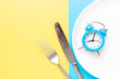 Blue alarm clock, fork, knife and empty plate on colored paper background. Intermittent fasting concept