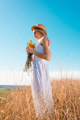 Wall Mural - side view of stylish woman in white dress and straw hat holding wildflowers against blue sky