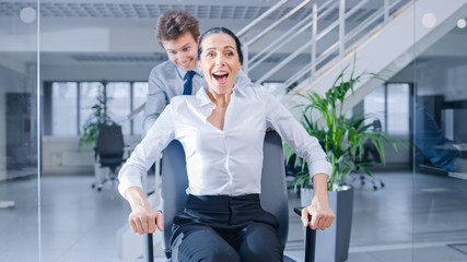 Poster - Cheerful Employee Pushes His Beautiful Female Colleague on a Chair Between Rows of Desks with Diverse Business People Working on Desktop Computers in Modern Office Space.