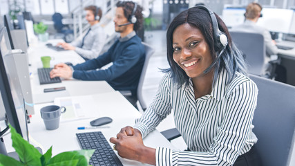 Poster - Beautiful Young Female Customer Service Operator Smiling for a Portrait at a Busy Modern Call Center with Diverse Multicultural Team of Office Specialists Wearing Headsets and Taking Calls. 