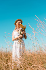 Wall Mural - selective focus of blonde woman in straw hat and white dress holding bouquet of wildflowers against blue sky