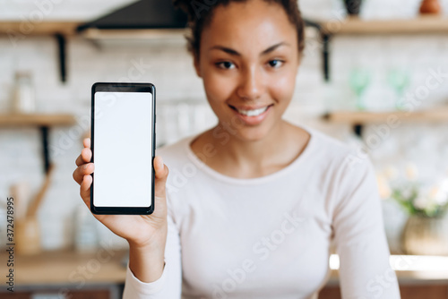 Cuadros en Lienzo Smiling, beautiful girl sitting in the kitchen, holding a phone in her hands wit