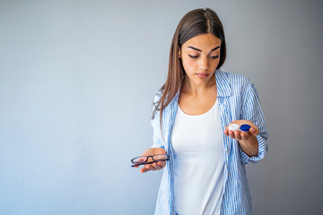 Wall Mural - woman holding contact lens case on hand and holding in her other hand a glasses on grey background., eyesight and eyecare concept. Young woman choosing between contact lenses or glasses