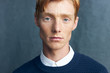 © monchak - portrait of a young handsome red-haired man with freckles on a dark background he is wearing a classic dark blue sweater on a white shirt  he looks at camera with serious face