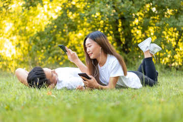 young asian couple lying down in the park listening music and relaxing with smartphones, boy and girl in love looking in each others eyes