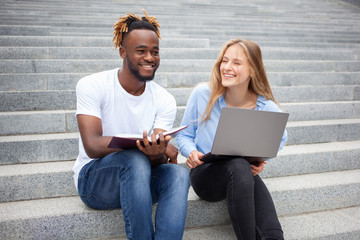 Education, learning and people concept - European young female and African American guy. Students sitting on stairs in the park, smiling and prepares for online exams.