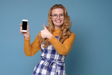 Mature woman housewife mother wearing kitchen apron showing a phone with empty screen. Studio shot on blue wall.