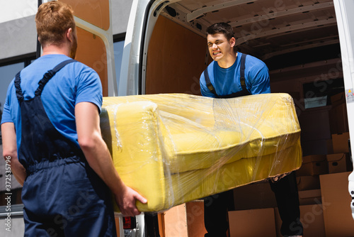 Selective focus of movers unloading sofa in stretch wrap in truck outdoors
