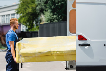 Wall Mural - Side view of loader in uniform holding couch in stretch wrap near truck with open doors on urban street