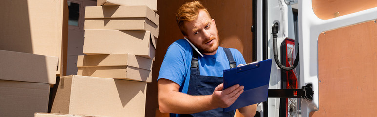 Poster - Panoramic shot of loader holding clipboard and talking on smartphone near packages in truck