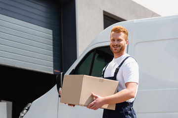 Wall Mural - Loader in overalls holding cardboard box and looking at camera beside truck on urban street