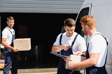 Wall Mural - Selective focus of loader with clipboard pointing with hand near colleague with cardboard box and truck on urban street